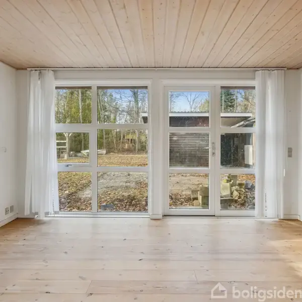 Et enkelt køkken med et træbord i midten, en hvid ovn ved væggen, og vinduer med udsigt til en skov. Døren fører ud til en terrasse.