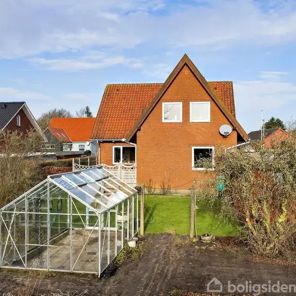 Huset er en rød murstensbygning med rødt tag, beliggende i en have med et drivhus i forgrunden. Omkringliggende huse og træer ses under en klar himmel.