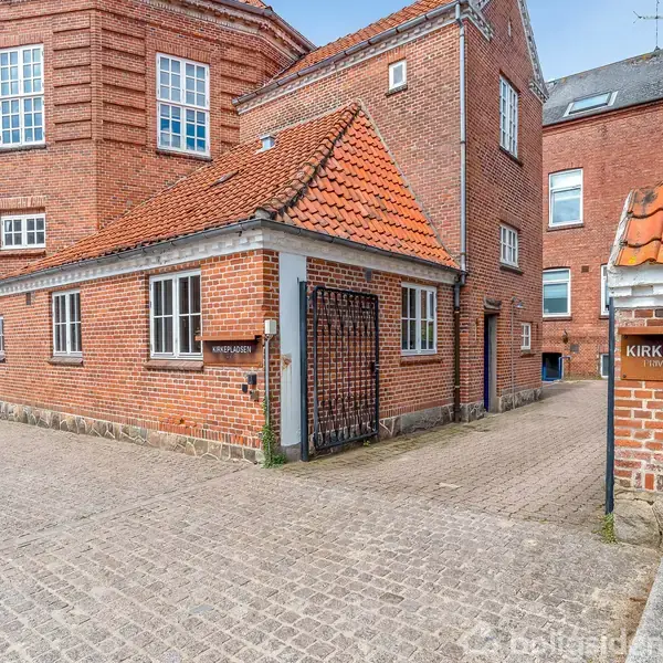 Bygning af røde mursten med tegltag, står stille. Skiltet ved indgangen viser "KIRKEPLADSEN - LUTHERSK MISSION", hvilket indikerer en religiøs kontekst i bymiljøet.