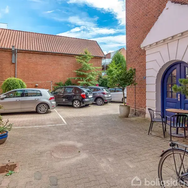 Parkerede biler holder stille i en gårdhave omgivet af murstensbygninger. Cykler og potteplanter står også i nærheden, mens himlen er klar og blå.