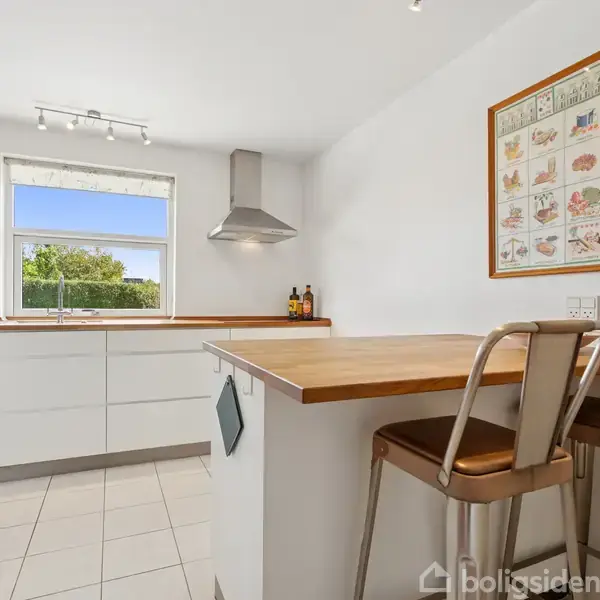 Køkkenbord med to høje stole står ved en lys køkkenø. Et vindue med udsigt til haven. Vægdekoration hænger ved siden af en emhætte over komfuret.