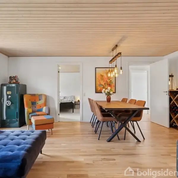 Blåt sofamøbel står i en lys stue med træloft og parketgulv. Spisebord med moderne stole er centralt placeret. Døre fører til andre rum, og et vinreol ses i højre side.