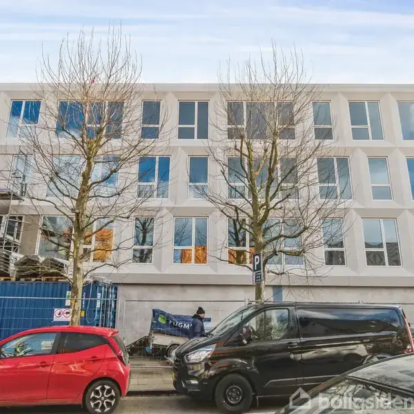 Bygning under opførelse med tre etager og store vinduer. Foran står to parkerede biler på en vej med træer. Området er afspærret med et hegn og byggeudstyr.