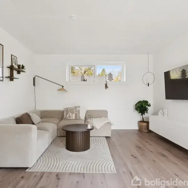 Stue indeholder en beige hjørnesofa med puder, en rund, brun sofabord og en vægmonteret lampe. TV hænger på væggen. Gulvet er lyst træ, og vinduer lukker lys ind.