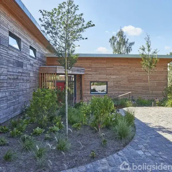 Moderne hus lavet af træ er omgivet af en velplejet have med grusbelagt indkørsel. Smalle vinduer pryder facaden, mens høje træer og buske tilføjer grønne omgivelser.