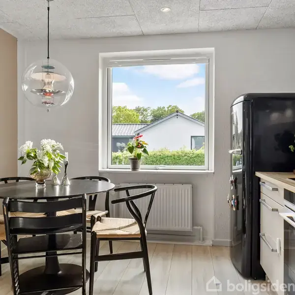 Et rundt spisebord med sorte stole står ved et vindue. En sort retro-køleskab står ved siden af et lyst køkkenbord. Planter og lysekrone pynter rummet.
