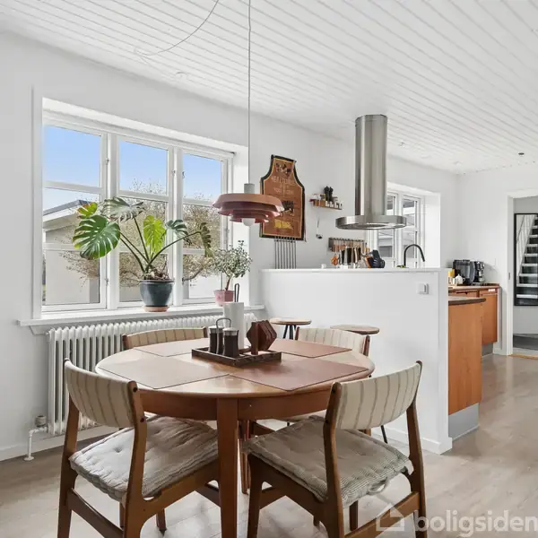 Et rundt spisebord med fire stole står i et lyst køkken med hvide vægge og store vinduer. På bordet ses dekorationer, og køkkenet er åbent mod et gangområde.