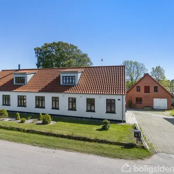 Hvid to-etagers bygning med rødt tegltag, står på en velplejet plæne ved en vej. I baggrunden ses en rød bygning og garage. Omgivet af træer og blå himmel.