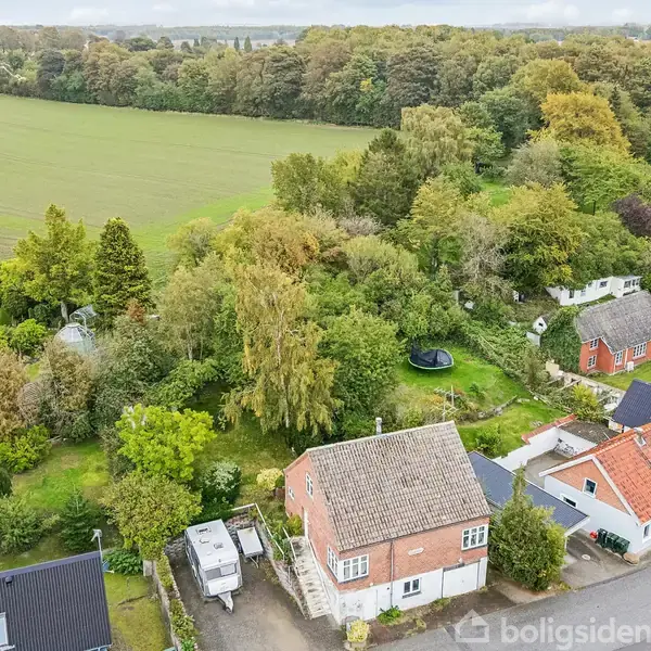 Rød murstenshus med gråt tag omgivet af frodige træer og haver. I forgrunden ses indkørsel og trailere. Omkringliggende område præget af flere huse og åbne marker i baggrunden.