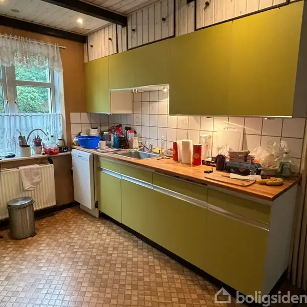 En køkken med gule skabe og en træbordplade, hvorpå der står køkkenredskaber og dåser. I baggrunden ses et vindue med hvide gardiner og en opvaskemaskine.
