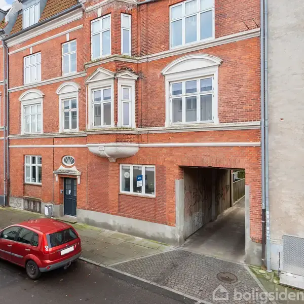 Rød murstensbygning med mange vinduer; bil parkeret foran. Smal passage fører igennem bygningen. Urban omgivelser med gade og nabohuse.