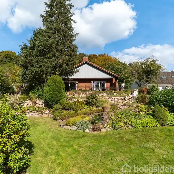 Huset står stille i en velplejet have med stenbede og grønne planter. Skyer pryder den blå himmel i baggrunden, der skaber en fredelig atmosfære.