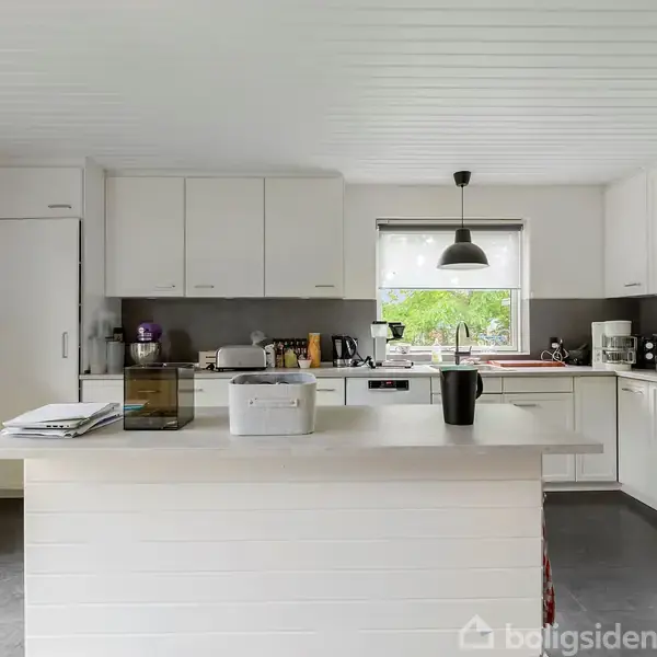 Køkken med hvidt interiør har en central ø med køkkenredskaber og papirbunker. Skabe og apparater omgiver rummet, mens et vindue viser træer udenfor. Stependently oven og kaffemaskine står til højre.