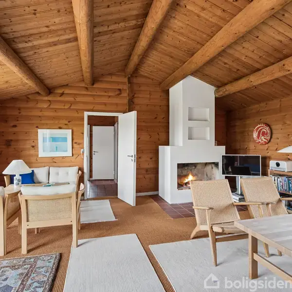 Stue med trævægge og loft, hvor en tændt brændeovn skaber hygge. Møbler inkluderer spisebord, stole og sofa. Vinduer åbner ud til lys omverden. Hylder og tæpper skaber hjemlig atmosfære.