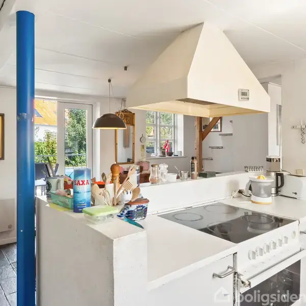 Køkken med hvidt komfur og emhætte, omgivet af hylder med køkkenartikler og et stort vindue med udsigt til haven. Et blåt rør står ved siden af døren.