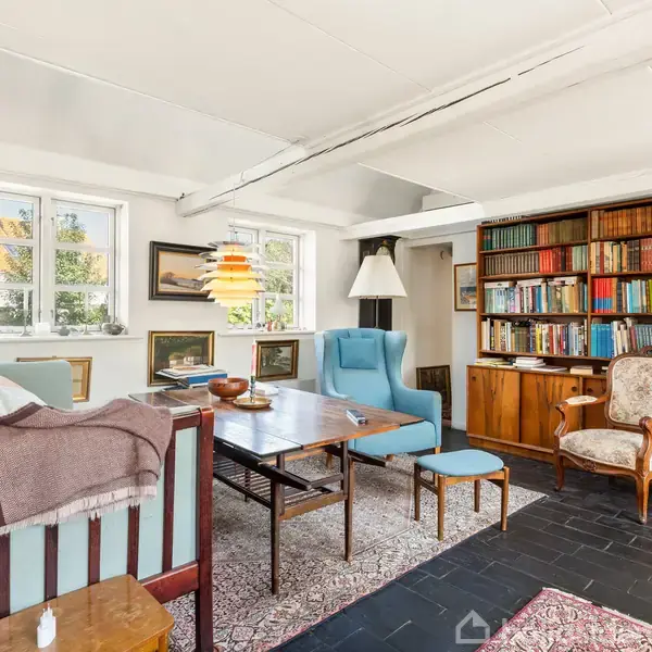 Et hyggeligt stueområde med en blå sofa, to blåt polstrede lænestole og et træbord. En bogreol fyldt med bøger står ved siden af en dekoreret lænestol, omgivet af tæpper.