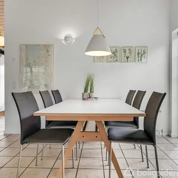 Et spisebord med sorte stole står i en lys spisestue. Bordet har en potteplante i midten, og der er kunst på væggen. Store vinduer giver udsigt til græsplænen.