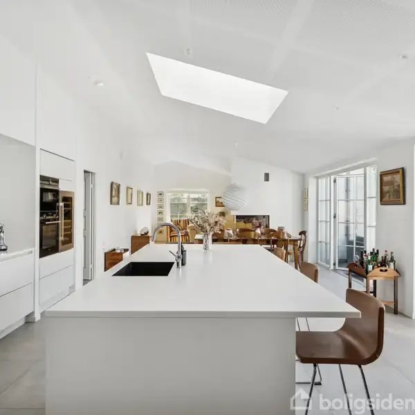 Køkkenalrum med stort, hvidt køkkenø i midten, omgivet af barstole. Et spisebord står i baggrunden ved store vinduer, der lukker sollys ind. Vægge dekoreret med malerier.