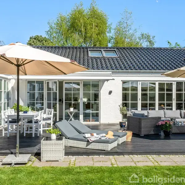 En terrasse med solsenge og parasoller står foran en hvid murstensvilla med store vinduer. Haven er grøn og omgivet af høje træer. Bord og stole er klar til brug.