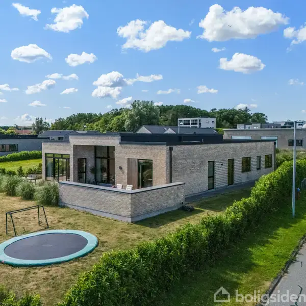 En moderne, lav murstensvilla med store vinduer står i en velholdt have. Ved siden af huset er der en vej, og i haven er der en nedgravet trampolin. Skyer ses på den blå himmel.