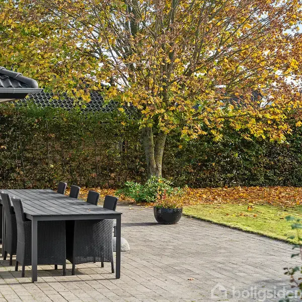 Lang havebord med sorte stole står på terrasse ved et murstenshus. En træ med gulnede blade samt græsplæne udgør haven i baggrunden.