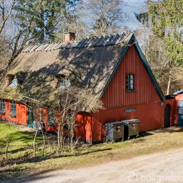 Et rødt landhus med stråtag står omgivet af træer. En grusvej fører forbi huset, mens skraldespande og en carport ses ved siden af.