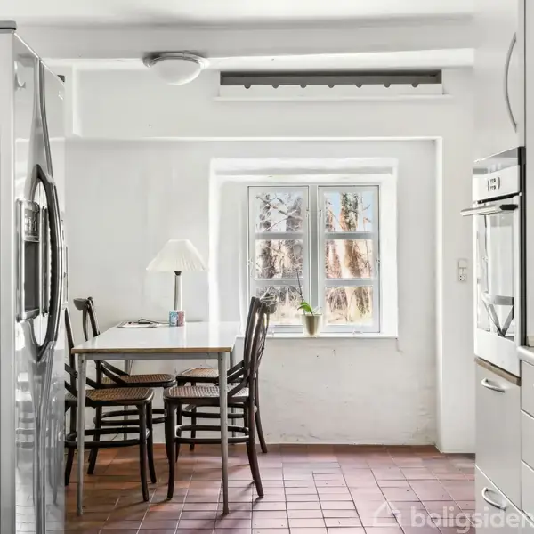 Køleskab står i køkkenet ved siden af et spisebord med tre stole, en lampe og et vindue med udsigt til træer. Rummet har hvide vægge og et flisegulv.