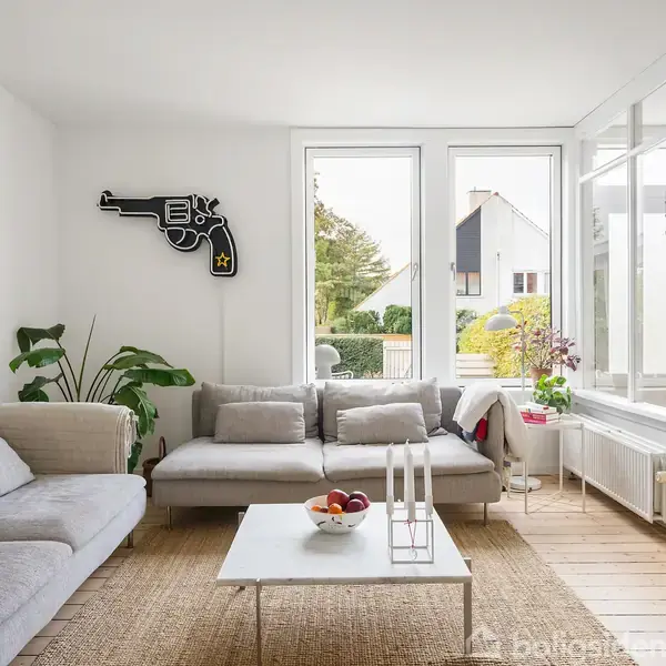 En lys stue har en grå hjørnesofa, placeret ved store vinduer. På væggen hænger en dekorativ revolverformet skulptur. Et lavt bord med lysestager og frugtskål står i midten.