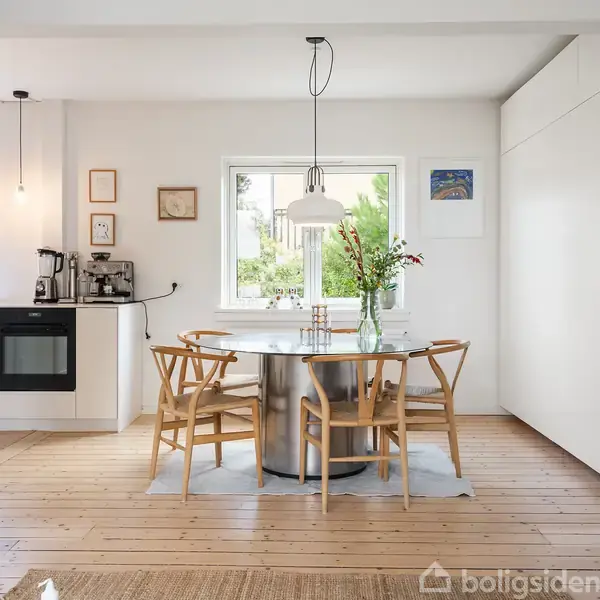 Rundt spisebord med træstole står i køkken-alrum, oplyst af hængelampe. Omkring bordet er lyse vægge med vindue og kunst. Kaffemaskine og ovn ses i baggrunden.
