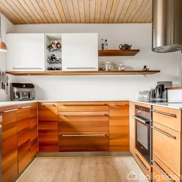 Et moderne køkken med træskabe, hvid bordplade, og indbygget ovn. Der er en række hylder med glas og køkkenredskaber, samt en stål-emhætte. Rummet er lyst og åbent.