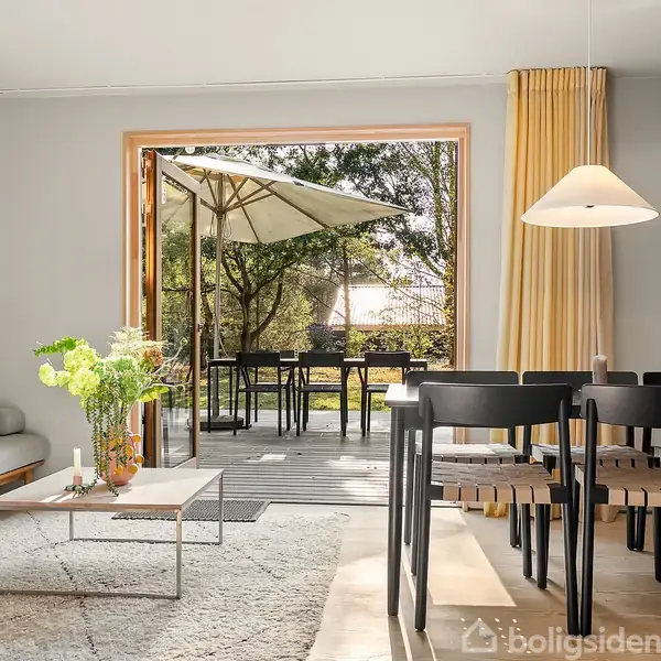 Sofa ved sofabord med vase og blomster står i stue. Spisebord med stole ved siden af. Åben terrassedør fører til terrasse med parasol. Sollys strømmer ind.