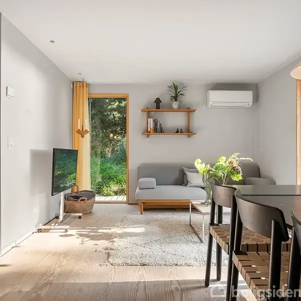 Stue med gråt hjørnesofa og tv på venstre side. Rundt spisebord med stole til højre. Store vinduer giver udsigt til grøn have. Hylde med planter og pyntegenstande.