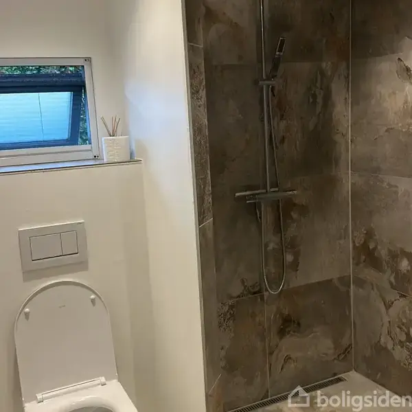 Toilet placeret på væg, står ved siden af en bruseniche med mørke fliser. En lille vinduesramme med udsigt findes ovenfor toilettet.