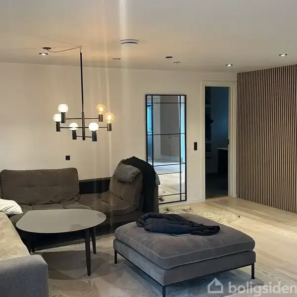 En stue med en grå hjørnesofa, sofabord og en stor puf. Der hænger en moderne lampe ovenover. I baggrunden ses en dør og trælister på væggen.