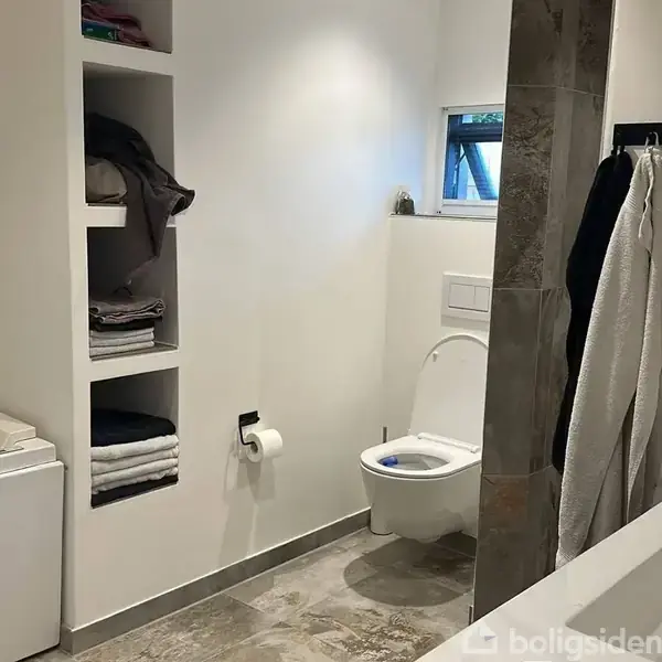 Toilet står stille i et moderne badeværelse med hvide vægge. Ved siden af er hylder med stablede håndklæder og tøj, og en toiletrulle hænger på væggen.