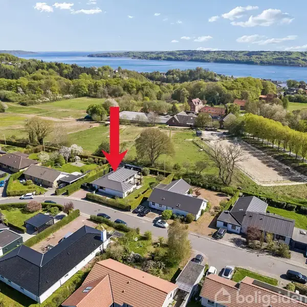 Et hus er markeret med en rød pil ovenfra, placeret i et villakvarter nær en bugt. Omkringliggende huse og grønne områder omkranser boligen.