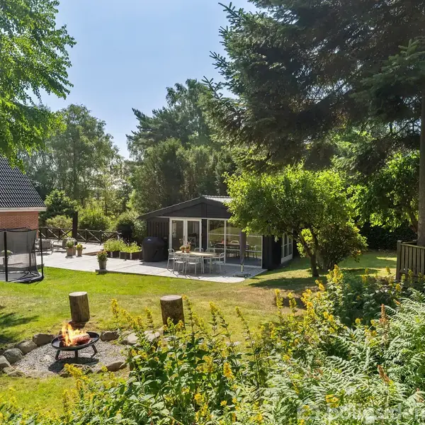 Et bål i en have omgivet af stubbede siddepladser, tæt på et moderne hus med terrasser og grønne træer omkring. Det er en solrig dag, og omgivelserne er frodige.