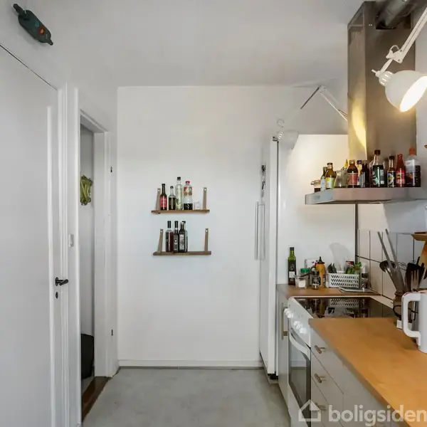 Køkken med hvide vægge og en træbordplade fyldt med køkkenredskaber og glas. Hylder med flasker dekorerer væggen. En hvid dør fører til et tilstødende rum.