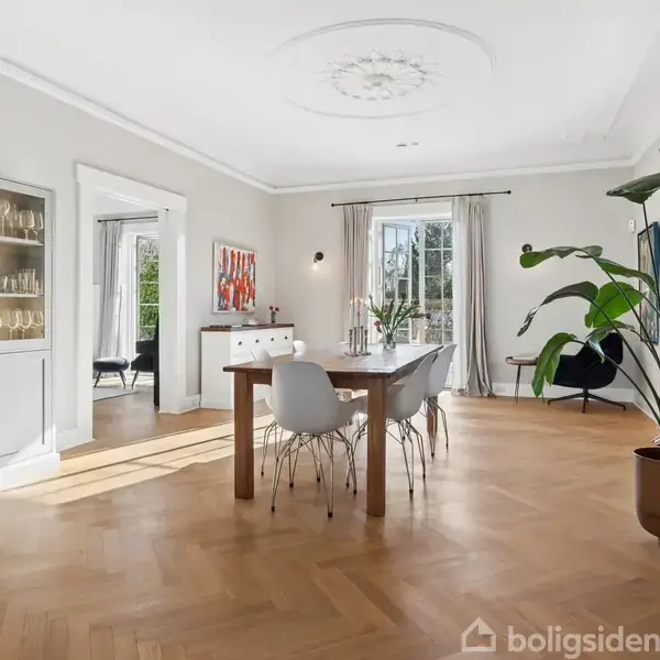 Spisebord med hvide stole står på et lyst trægulv i en rummelig spisestue. Store vinduer og dør med lyse gardiner, hvide skabe og en grøn plante skaber en elegant atmosfære.