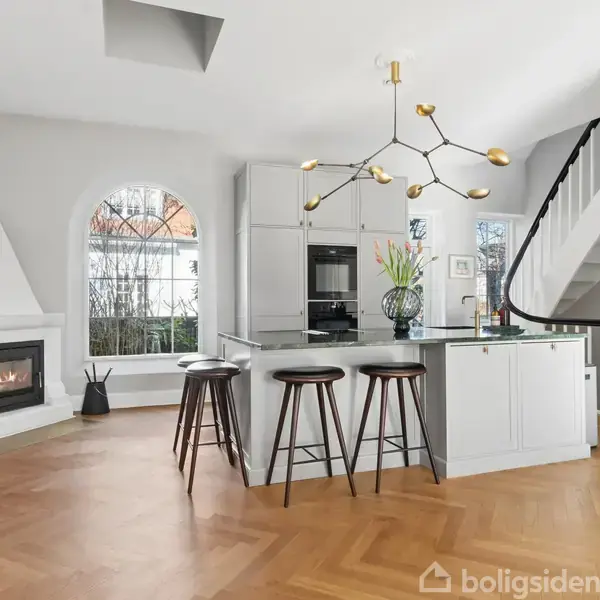 Køkken med ø og tre barstole, moderne lysekrone, pejs til venstre, trappe til højre, stor bueformet vindue bagved, minimalistisk indretning, lyst trægulv, vase med blomster på bordet.
