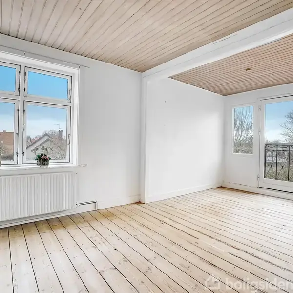 Et lyst rum står tomt med trægulve, lyse vægge og loft. Store vinduer og en glasdør giver udsigt til træer udenfor. To planter står på vindueskarmen.