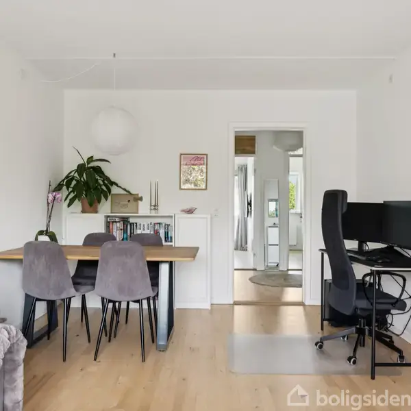 Spisebord med fire stole står ved en væg med planter og kunst, mens et skrivebord med computer er placeret i et hjemligt rum med lyse trægulve og lyse vægge.