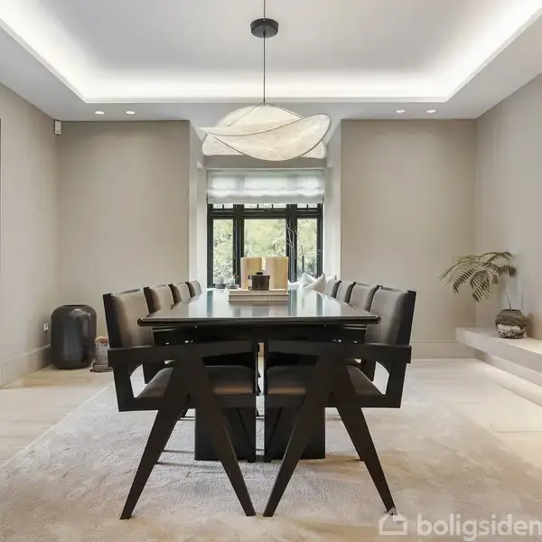 Et spisebord med stole står i midten af et elegant spisestue. Lampe hænger over bordet, omgivet af dæmpet belysning. Moderne indretning og store vinduer skaber en lys atmosfære.