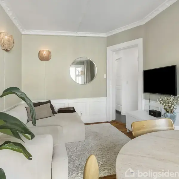 En stue med en beige sofa, der står mod en væg. Et rundt spejl hænger over sofaen. Fladskærms-tv på væggen til højre, planter og rundt bord nær sofaen.