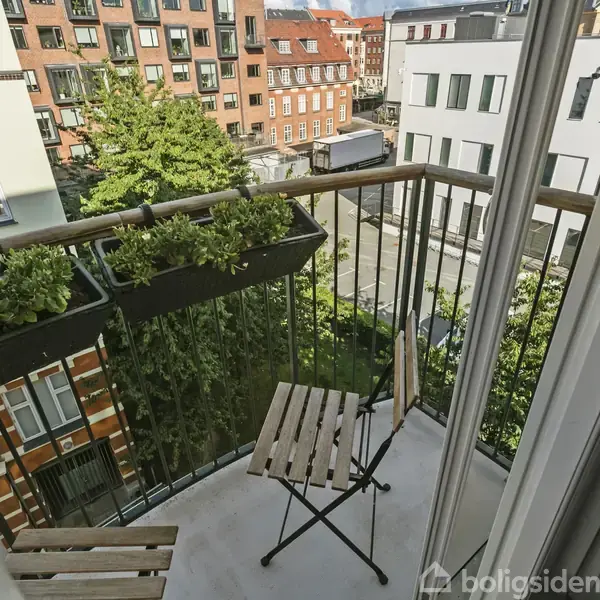 Balkonen har to træstole og blomsterkasser med planter. Den vender ud mod en byudsigt med bygninger og grønne træer under en blå himmel.