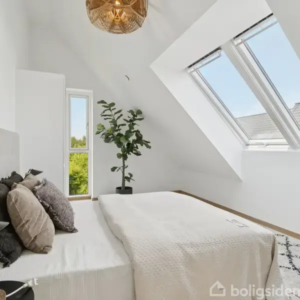 Et soveværelse med en seng dækket af puder og tæpper. Ved siden af står en lampe og en plante. Rummet har skråvægge med store vinduer og en hængende lampe.