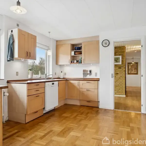 Køkken med træskabe og parketgulv. Opvaskemaskine og ovn er indbygget. Rummets vægge har store vinduer, og en dør åbner til en gang med synlig murstensvæg. En vægur hænger over døren.