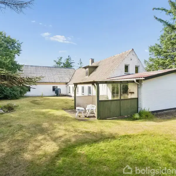 Et hvidt hus med en lille veranda står stille midt i en græsplæne omgivet af træer. To hvide stole er placeret foran verandaen.