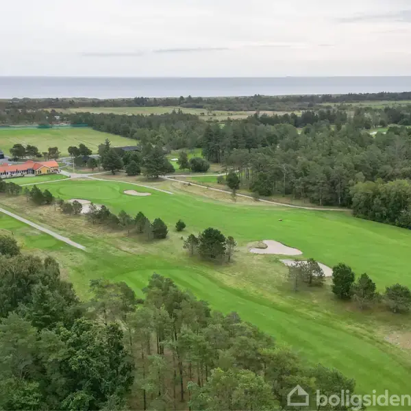 Golfbane strækker sig over et grønt landskab nær skov, med få træer på græsarealet. I baggrunden kan man se havet og en bygning med teksten "FUR IDRÆTSCENTER".