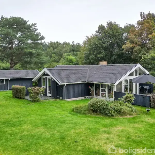 Et sortmalet træhus med store vinduer står på en grøn græsplæne omgivet af buske og træer. Ved siden af ligger et mindre skur. Skyfri himmel i baggrunden.